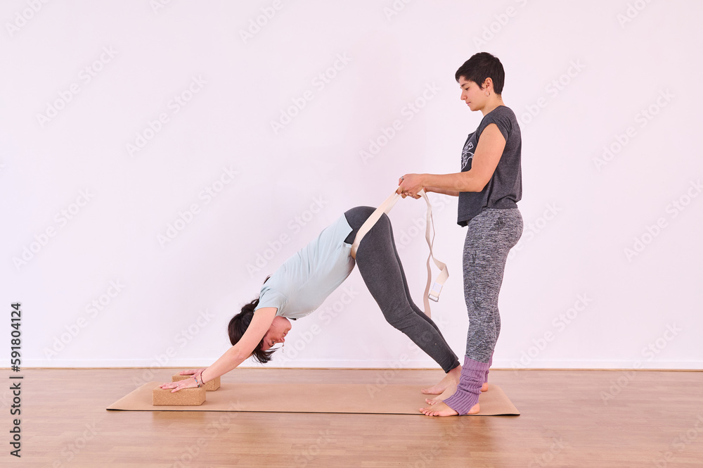 Two girls practicing yoga, one holding the other with a white strap around the waist