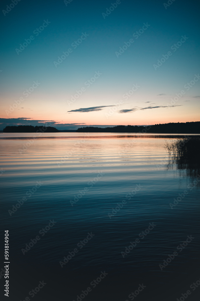 Vertical of a tranquil lake reflecting a rocky shore covered with plants at sunset