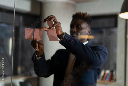 Portrait of successful businessman in office. Young African man witing ideas on colorful stickers on glass wall.