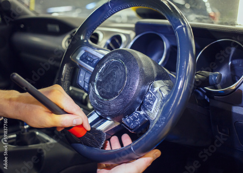 Car detailing and the concept of car interior care. A young man cleans the steering wheel of a modern car using a special brush. Dry cleaning of the car interior, close-up, selective focus.