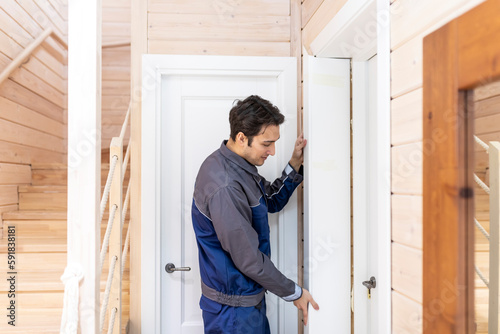 Installation of a wooden door. Portrait of young handyman in blue uniform installing door. Professional repair service and maintenance concept