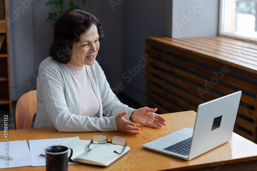 Happy middle aged senior woman sit with laptop talk on video call with friends family. Laughing mature old senior grandmother having fun talking speaking with grown up children online