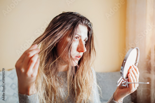 Young annoyed woman is upset about dirty oily and greasy hair looking in mirror at home. Health treatment