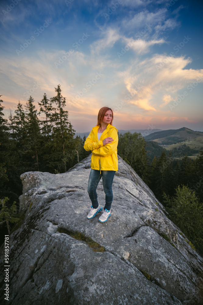 The woman in yellow jacket on the boulder, the top of rock. Sunset summer day. Vertical photo. Active lifestyle recreation, hiking and travel. Beautiful view of the Carpathians, Ukraine.