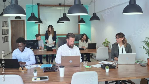 Real time high angle of multiracial colleagues in formal outfits sitting in open space office at tables with laptops while communicating and discussing details of business project