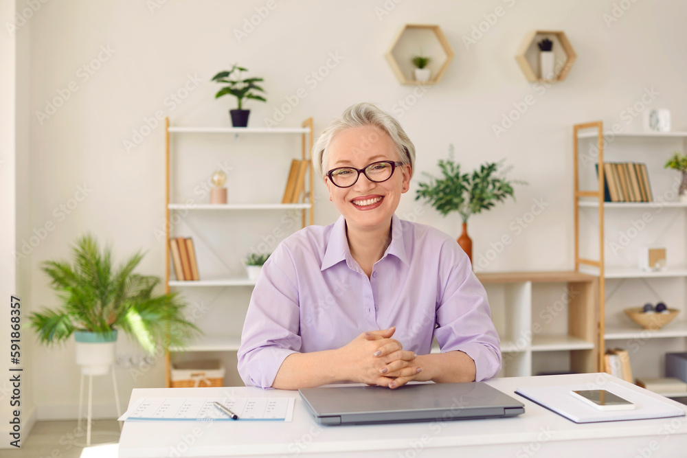 Portrait of a happy elegant businesswoman in her office. Cheerful, smiling, good looking mature woman in a shirt and eyeglasses with short white hair sitting at her working desk with a laptop Portrait of a happy elegant businesswoman in her office. Cheerful, smiling, good looking mature woman in a shirt and eyeglasses with short white hair sitting at her working desk with a laptop