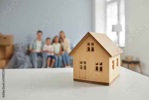 Wooden small house close-up on table and family sitting on couch in cozy room moving in new house with boxes on background. Relocation, mortgage, buying a property, moving, owning real estate concept.