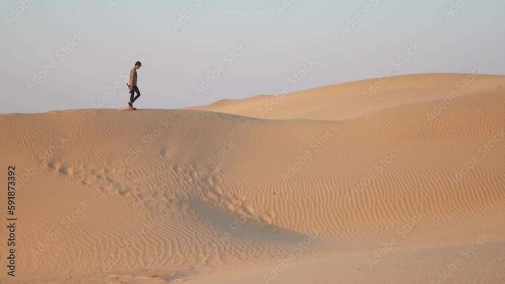 Indian man walking on the top of the sand dunes of the Thar desert in Jaisalmer, Rajasthan, India. Tourist enjoying the desert sand while walking on the top of the sand dune. Desert tourism background Indian man walking on the top of the sand dunes of the Thar desert in Jaisalmer, Rajasthan, India. Tourist enjoying the desert sand while walking on the top of the sand dune. Desert tourism background