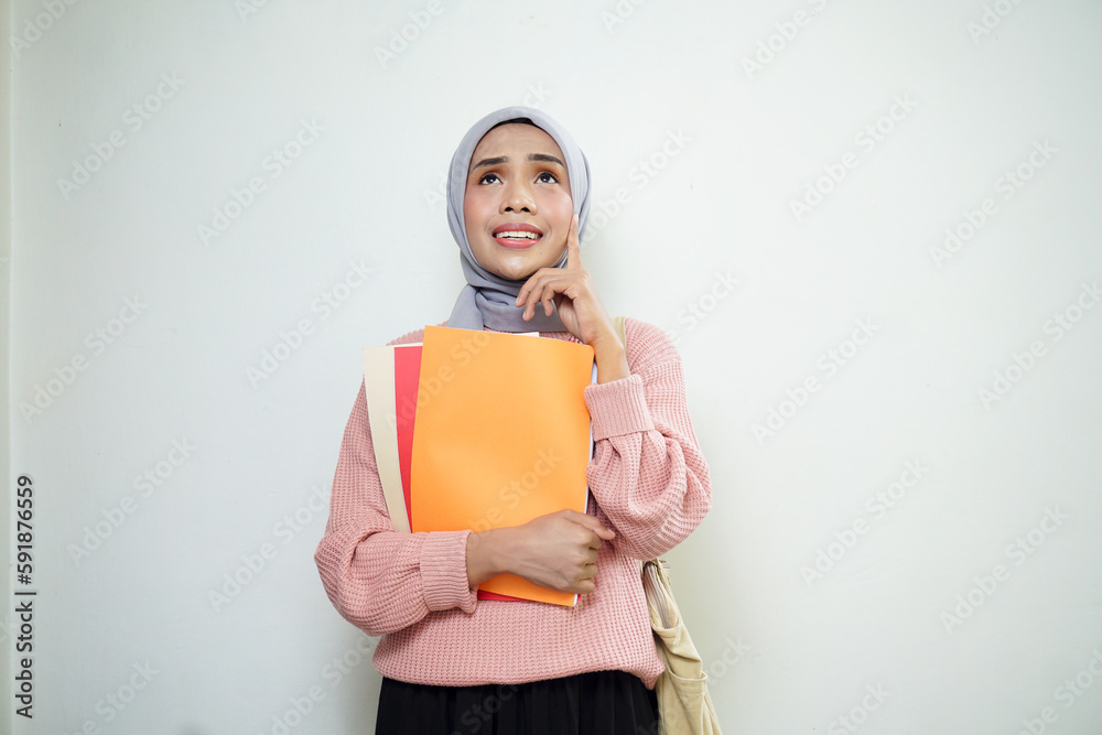 Smiling Asian Muslim female student in pink sweater with medium bag, holding book and thinking isolated on white background. back to school concept Smiling Asian Muslim female student in pink sweater with medium bag, holding book and thinking isolated on white background. back to school concept