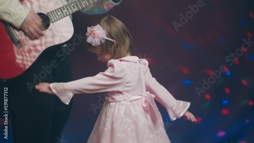 Little girl on stage in pink vintage dress, she dances, her father plays acoustic guitar. Color music is shining and smoke is billowing. Father Day. Performance on stage. Hobbies and creativity