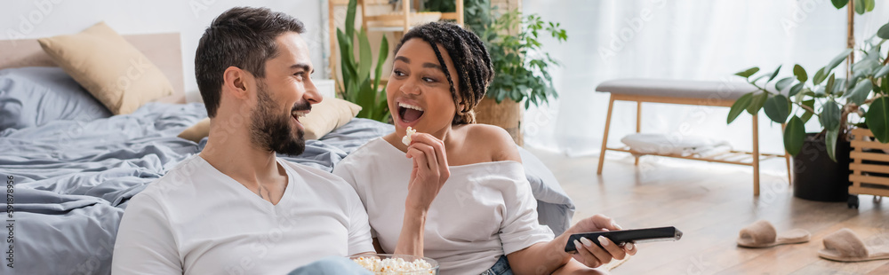 excited bearded man feeding african american woman with popcorn while watching tv in bedroom at home, banner. excited bearded man feeding african american woman with popcorn while watching tv in bedroom at home, banner.