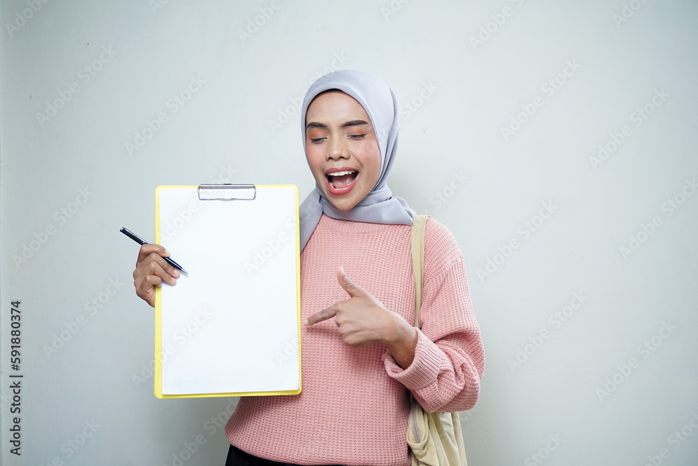 Smiling Asian Muslim female student in pink sweater with bag, holding board and pointing at empty board isolated on white background. back to school concept Smiling Asian Muslim female student in pink sweater with bag, holding board and pointing at empty board isolated on white background. back to school concept