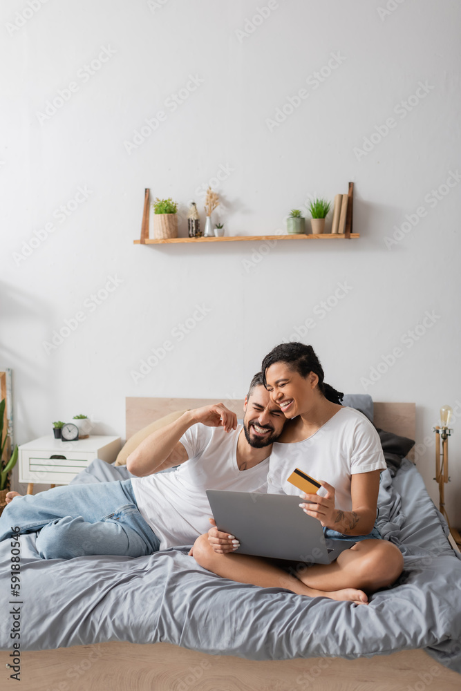 cheerful multiethnic couple with laptop and credit card laughing during online shopping on bed at home. cheerful multiethnic couple with laptop and credit card laughing during online shopping on bed at home.