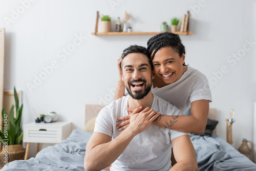 overjoyed multiethnic couple in white t-shirts laughing at camera in bedroom at home.