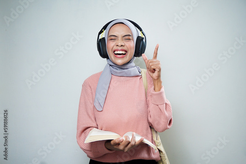 Smiling Asian Muslim female student in pink sweater with bag while listening music excitedly, set and reading book isolated on white background. back to school concept