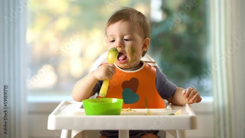The baby with food stained face trying to eat himself with a spoon sitting in baby chair