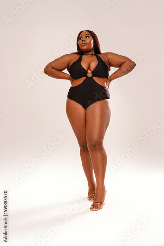 Woman posing on light gray studio background, wearing black fashionable swimwear. Body positive, conscious concept. Afro hairstyle.