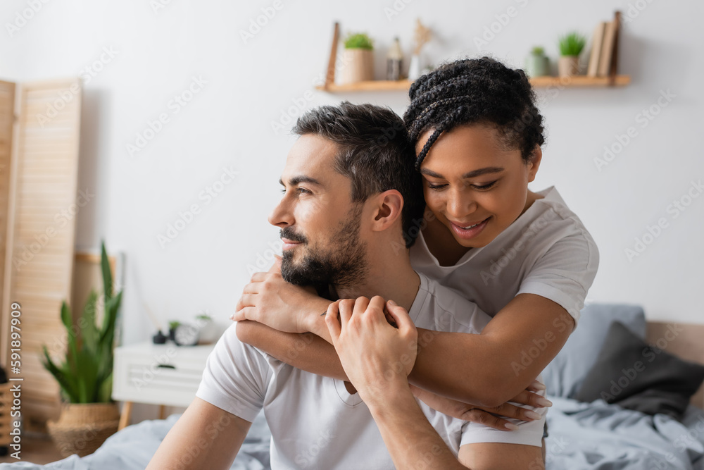 Fototapeta premium happy african american woman embracing dreamy bearded man looking away in bedroom at home.