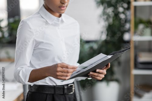 partial view of young african american manager in white blouse holding folder with document while standing in office.