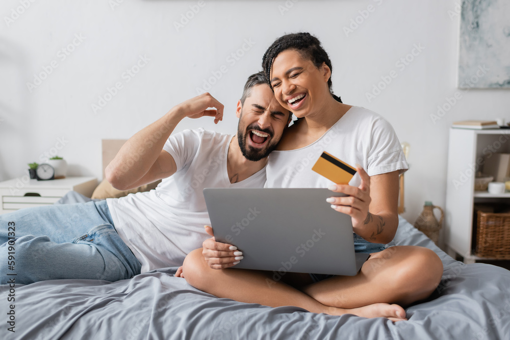 overjoyed interracial couple laughing with closed eyes near laptop and credit card on bed at home.