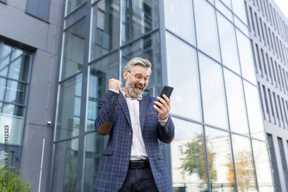 A senior man in a suit stands near an office center and is happy. Shows a victory gesture with his hand and shouts. He looks at his phone, reads a message, a letter, got a job, position, promotion. A senior man in a suit stands near an office center and is happy. Shows a victory gesture with his hand and shouts. He looks at his phone, reads a message, a letter, got a job, position, promotion.