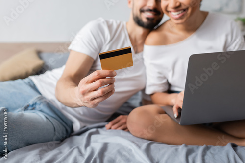 cropped view of blurred man holding credit card near smiling african american woman with laptop in bedroom at home.