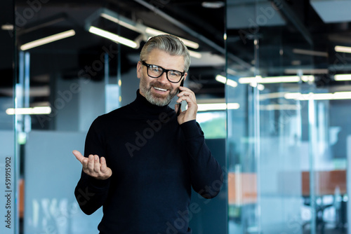 Portrait of an older gray-haired businessman standing in the office and smiling talking on the phone. Business conversation with partners. agreement on a meeting, resolution of issues.