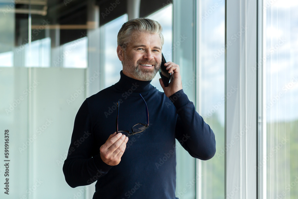 Φωτογραφία Portrait of senior male businessman standing in office near window, holding glasses in hand and smiling talking on phone, looking at camera