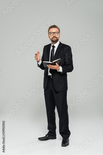 full length of well dressed businessman in glasses and formal wear holding notebook on grey.