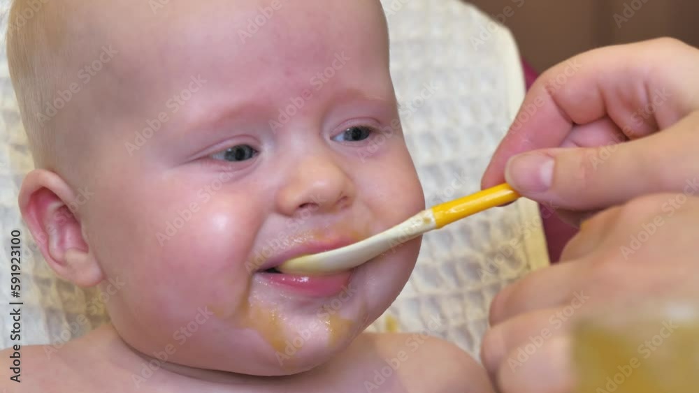 Mom feeding little boy with broccoli puree. Child at the age of six months eats broccoli while sitting on a baby chair. Slow motion shot. Mom feeding little boy with broccoli puree. Child at the age of six months eats broccoli while sitting on a baby chair. Slow motion shot.
