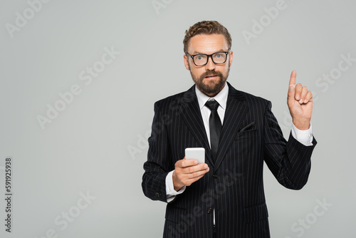 bearded businessman in glasses and suit holding smartphone and pointing with finger isolated on grey .