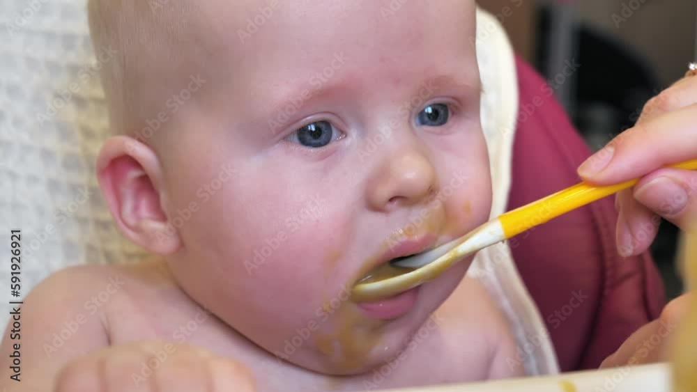 Mom feeding little boy with broccoli puree. Child at the age of six months eats broccoli while sitting on a baby chair. Slow motion shot. Mom feeding little boy with broccoli puree. Child at the age of six months eats broccoli while sitting on a baby chair. Slow motion shot.