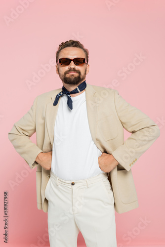 stylish french man in neck scarf and sunglasses holding posing with hands on hips isolated on pink.