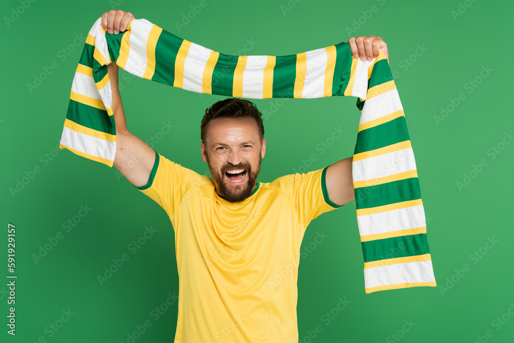 Φωτογραφία excited soccer fan in yellow t-shirt holding striped scarf and looking at camera isolated on green
