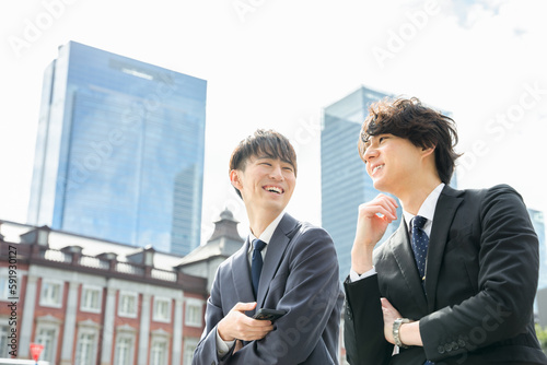 東京駅の見えるビジネス街で歩きながら仕事の話をするスーツ姿で笑顔の20代〜30代の2人の男性 photo