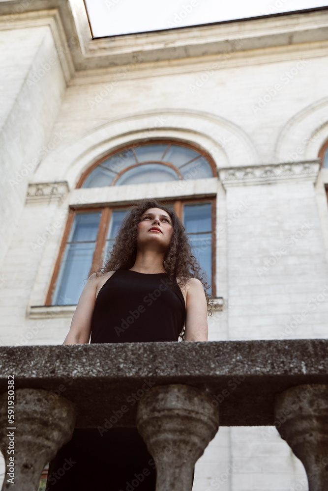 A beautiful portrait of a curly brunette girl in a black dress and makeup, poses gorgeous on balcony, looking up. Beauty portrait. 
