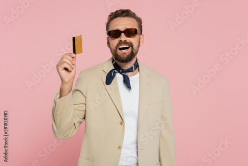 excited man in stylish sunglasses and neck scarf holding credit card isolated on pink.