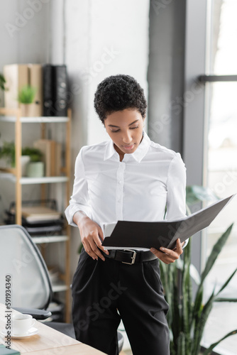 stylish african american businesswoman in white blouse and black pants standing with folder in office.