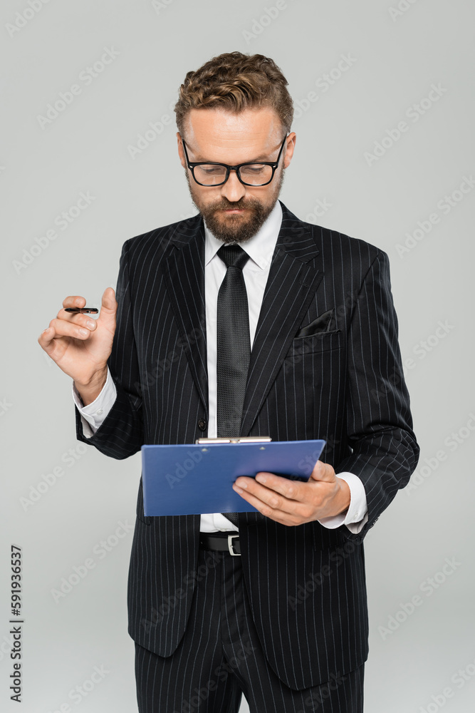 well dressed businessman in glasses and formal wear holding clipboard with pen isolated on grey.
