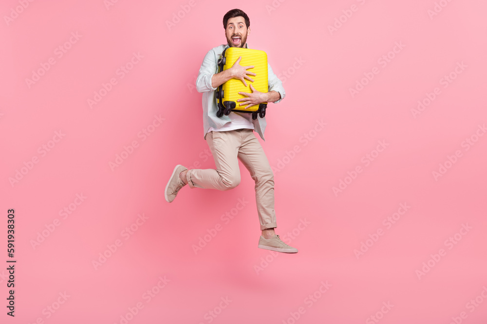 Full length photo of excited funny guy dressed teal shirt jumping high embracing suitcase isolated pink color background