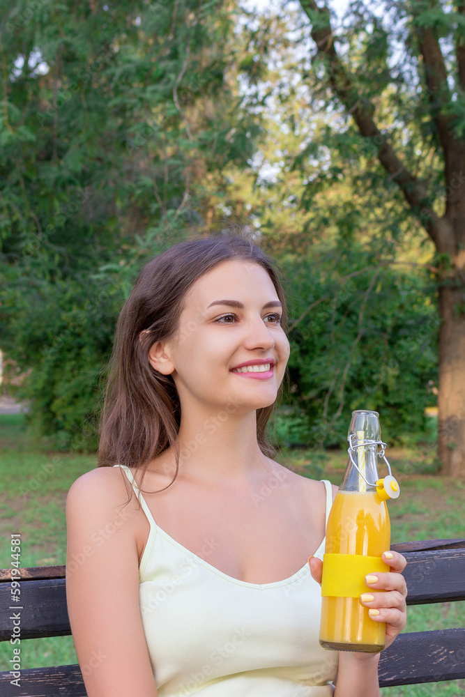 Attractive young woman is sitting on the bench in the park and holding a bottle of fresh juice with a straw Attractive young woman is sitting on the bench in the park and holding a bottle of fresh juice with a straw