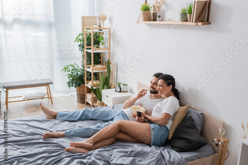 Wallpaper Mural full length of barefoot interracial couple eating popcorn and watching tv in modern spacious bedroom. Torontodigital.ca