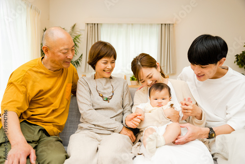自宅のリビングに0歳の赤ちゃんを囲って座る笑顔で幸せなシニアの祖父母夫婦と両親の3世帯の5人家族