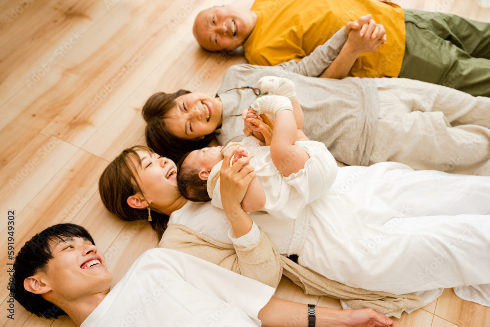 床に仰向けに寝そべり手を繋ぐ0歳の赤ちゃん含む3世帯5人の幸せで笑顔の家族