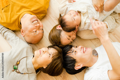 床で顔を近づけて仰向けで寝そべる0歳の赤ちゃんと両親とシニア世代の祖父母の3世代の家族5人