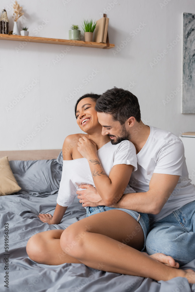 bearded man in white t-shirt hugging carefree african american woman sitting on bed at home. bearded man in white t-shirt hugging carefree african american woman sitting on bed at home.