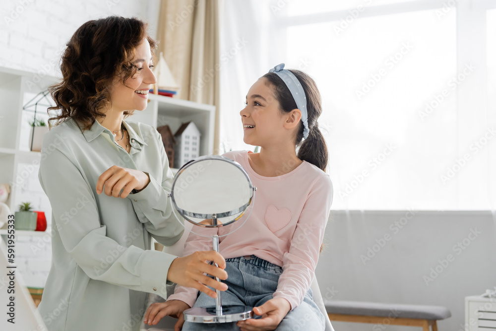 Obraz premium Smiling speech therapist holding mirror and looking at pupil in consulting room.