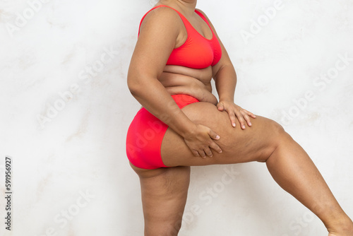 Overweight woman standing sideways massage raised thigh by hands, free copy space, white background. Bare woman in red underwear with cellulite body. Plus size people, feminism, self acceptance.