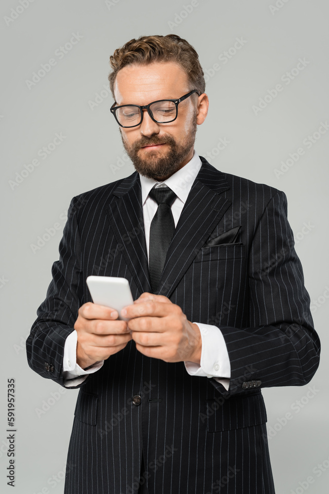 bearded businessman in glasses and suit typing on smartphone isolated on grey . bearded businessman in glasses and suit typing on smartphone isolated on grey .