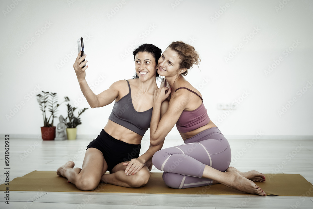 Two fitness women friend sitting on mat together and taking selfie on phone indoors Two fitness women friend sitting on mat together and taking selfie on phone indoors